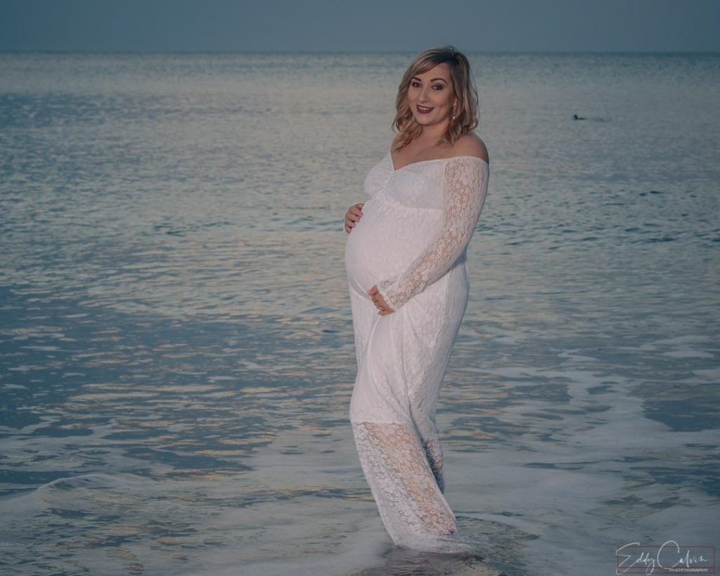 A pregnant woman in a white dress standing in the ocean, captured by a Northern Ireland family photographer.