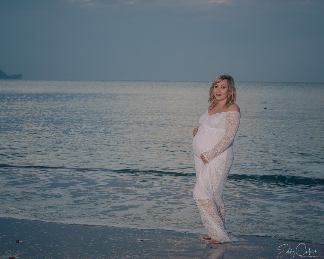 A pregnant woman standing on the beach at sunset, captured by a family photographer.