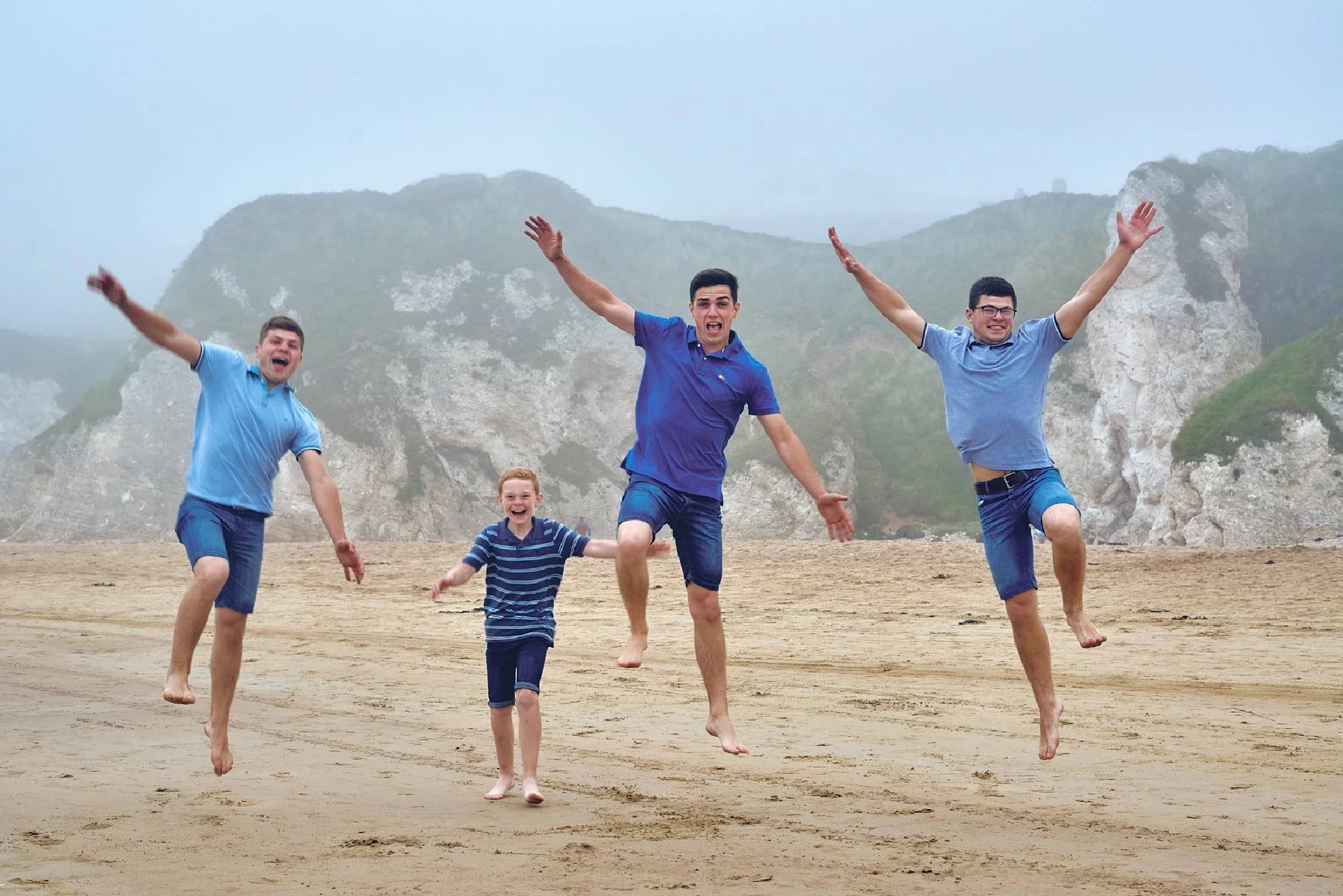 Family Photographer Four people performing jumping poses on a foggy beach with cliffs in the background.