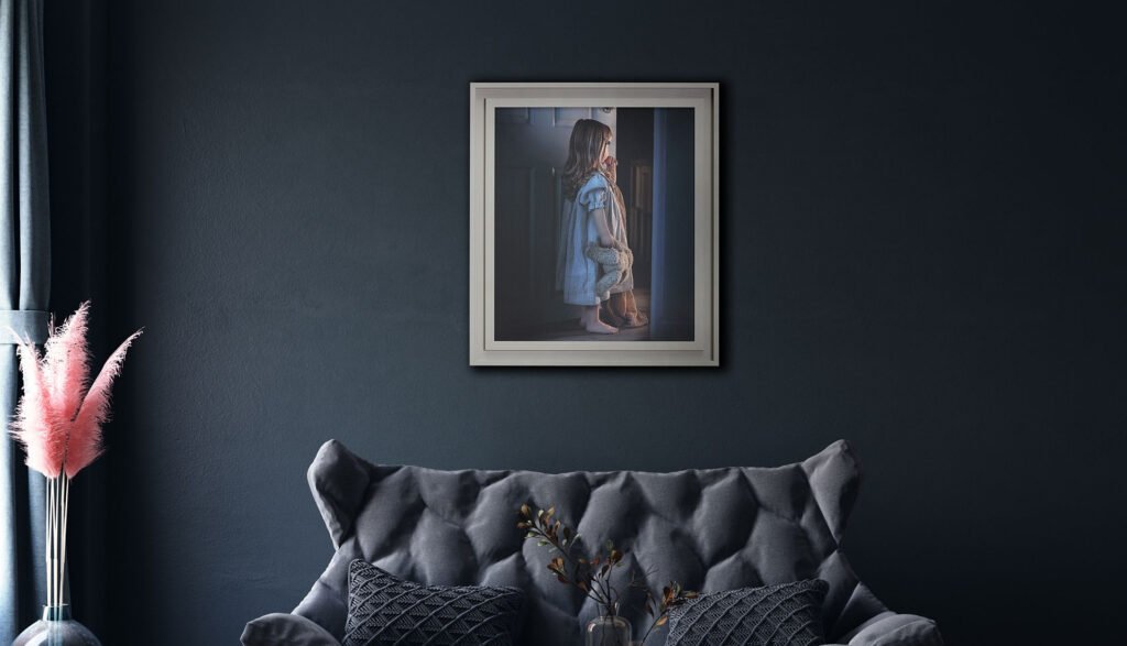 Family Photographer A framed photograph of a woman standing by a window is mounted on a dark-colored wall above a sofa with patterned cushions. a vase with pink and brown-toned feathers is placed to the left.